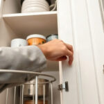 A hand in a gray sleeve reaching into an open white kitchen cabinet holding bowls and containers.